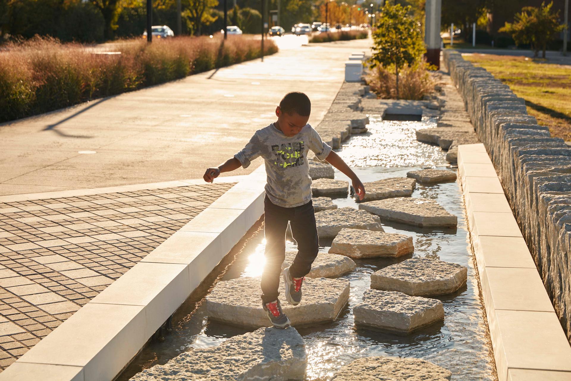 A kid plays along designed rocks in a water feature along Town Branch Commons. 