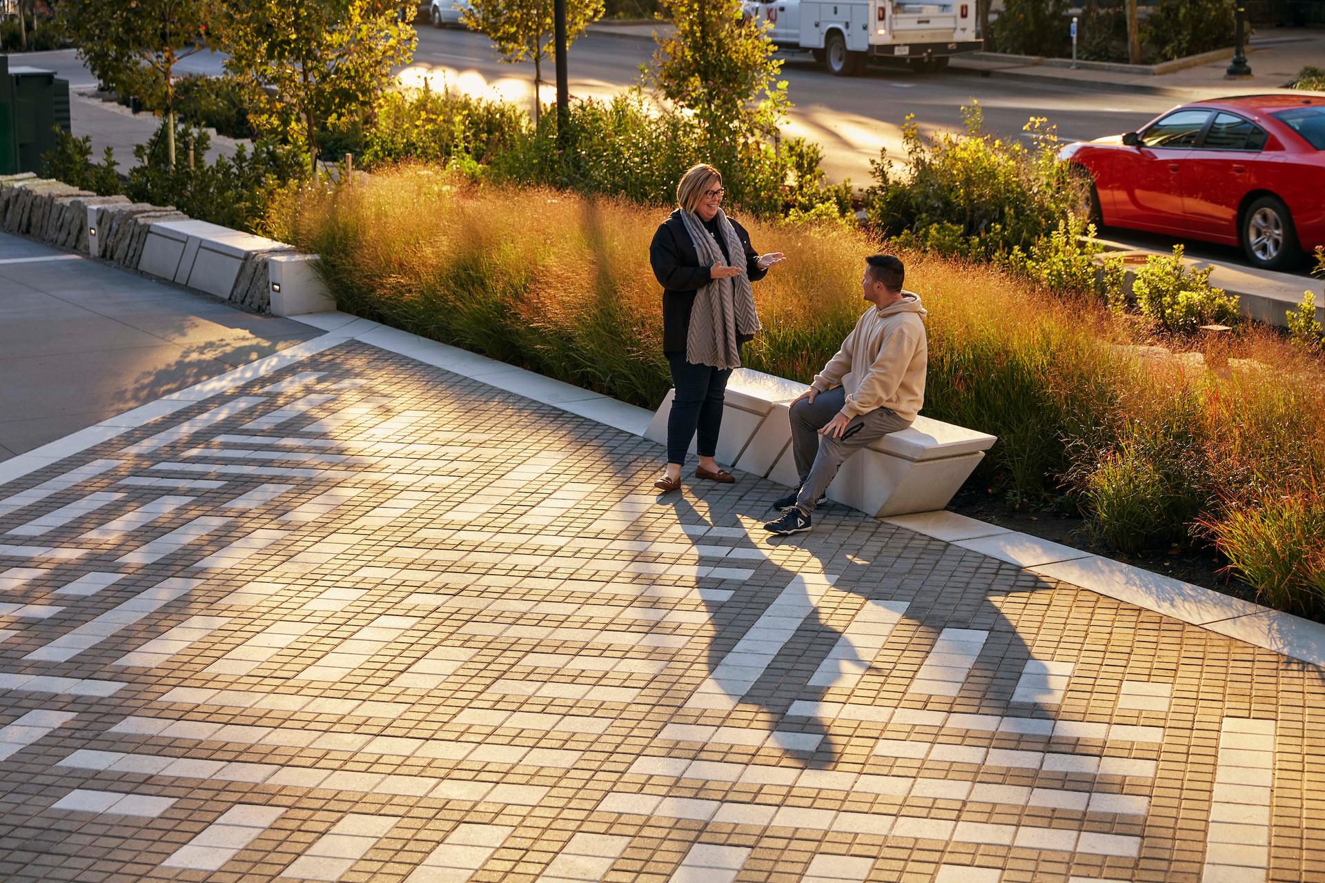 Two people stop to talk along Town Branch Commons. The sidewalk is wide with a white design of pavers. 