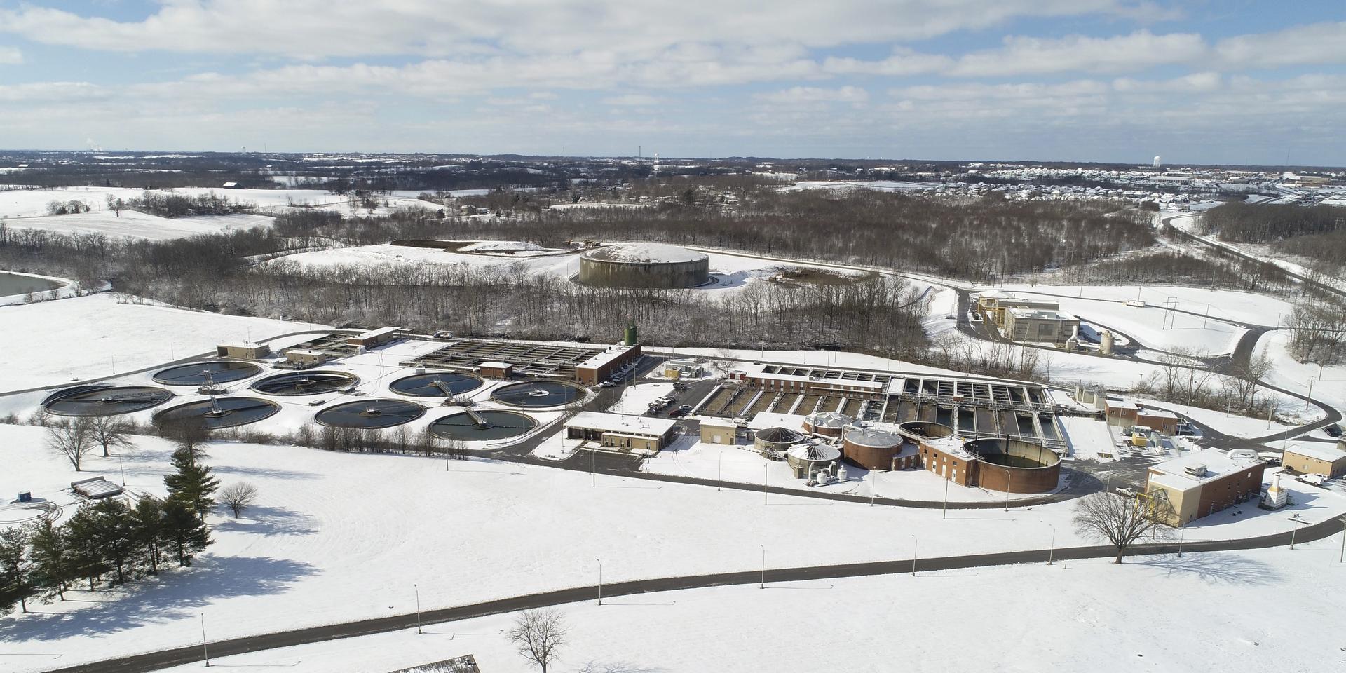 a overhead view from a drone of the West Hickman Wastewater Treatment Plant in the snow. There are trees surrounding the buildings and a blue sky.
