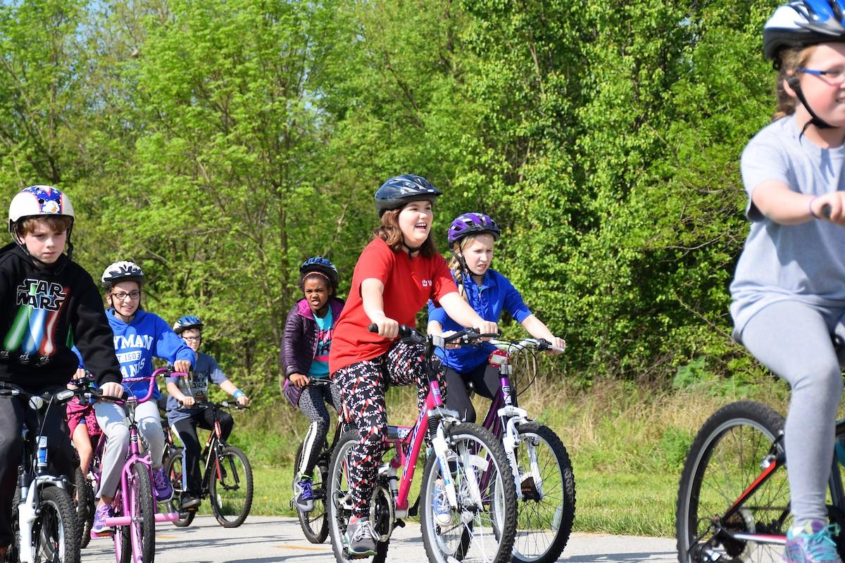 A group of children riding bikes on the Legacy Trail on a sunny day. A girl near the front has a big smile on her face. 
